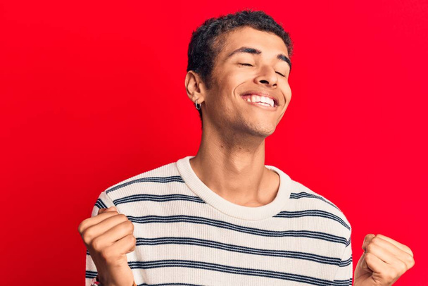 Junger afrikanisch-amerikanischer Mann in lässiger Kleidung sehr glücklich und aufgeregt dabei Siegergeste mit erhobenen Armen, lächelnd und schreiend nach Erfolg. Festkonzept.  - Foto, Bild
