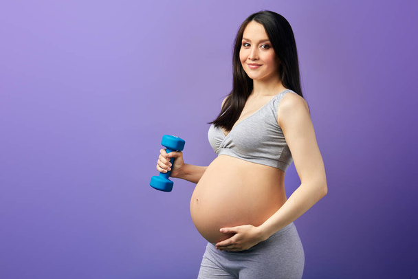 Happy pregnant mom exercising at studio with dumbbells on violet background - Fotografie, Obrázek