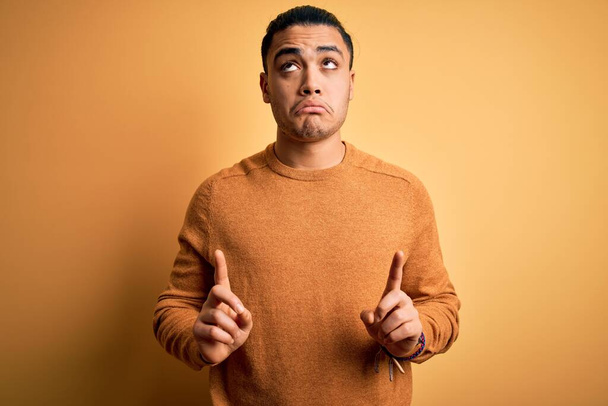 Young brazilian man wearing casual sweater standing over isolated yellow background Pointing up looking sad and upset, indicating direction with fingers, unhappy and depressed. - Φωτογραφία, εικόνα