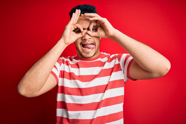 Jonge knappe man draagt casual gestreepte t-shirt en bril over geïsoleerde rode achtergrond doet ok gebaar als verrekijker steken tong uit, ogen kijken door vingers. Gekke uitdrukking. - Foto, afbeelding
