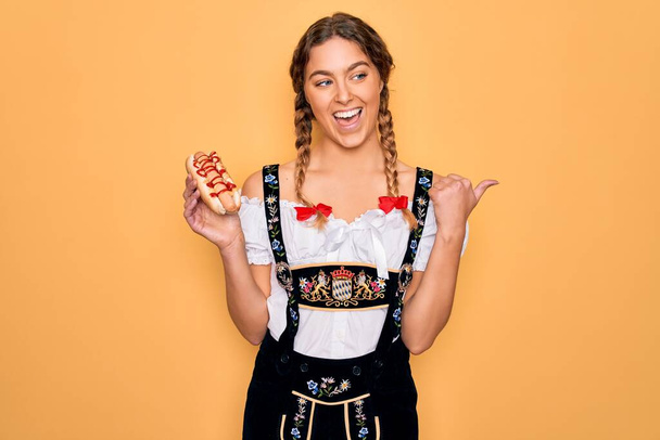 Mavi gözlü, güzel, sarışın Alman kadın. Oktoberfest elbisesi giyiyor. Sosisli yiyor, parmaklarıyla işaret ediyor ve gülümsüyor. - Fotoğraf, Görsel