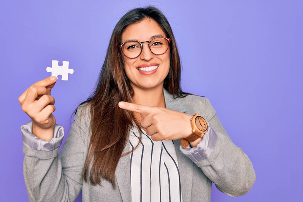 Young hispanic business woman holding puzzle peace as cooperation and union on work very happy pointing with hand and finger - Fotó, kép