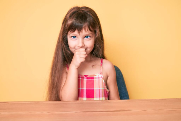 Kleines kaukasisches Mädchen mit langen Haaren in lässiger Kleidung sitzt auf dem Tisch und fühlt sich unwohl und hustet als Symptom für Erkältung oder Bronchitis. Gesundheitskonzept.  - Foto, Bild