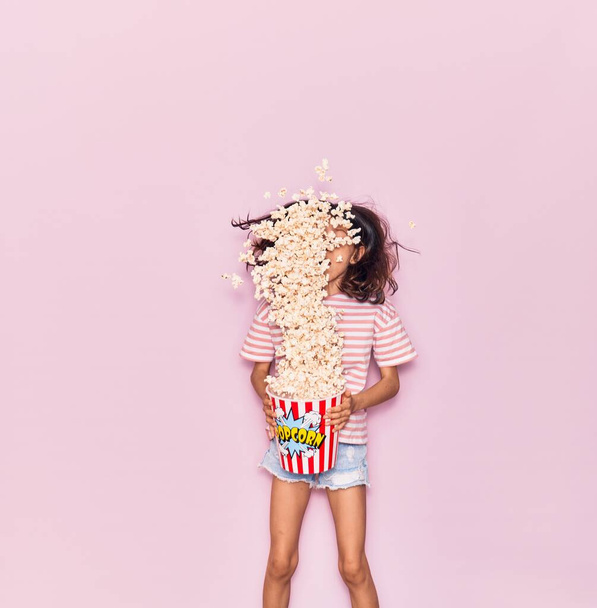 Adorable hispanic child girl surprised with open mouth holding bucket of popcorn jumping over isolated pink background - 写真・画像