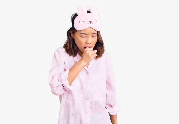 Young beautiful chinese girl wearing sleep mask and pajama feeling unwell and coughing as symptom for cold or bronchitis. health care concept.  - Photo, Image