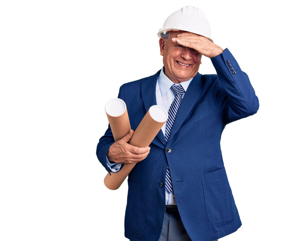 Älterer, gutaussehender grauhaariger Mann mit Architektenhut, der die Baupläne gestresst und frustriert von der Hand auf den Kopf hält, überrascht und wütend im Gesicht  - Foto, Bild
