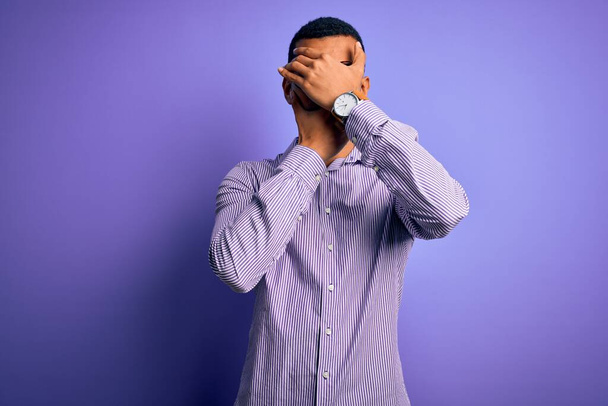 Knappe Afrikaans-Amerikaanse man met gestreept shirt en bril over paarse achtergrond Ogen en mond bedekken met handen, verrast en geschokt. Emotie verbergen - Foto, afbeelding