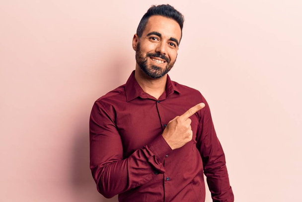 Junger hispanischer Mann in lässiger Kleidung lächelt fröhlich und zeigt mit der Hand und dem Finger zur Seite  - Foto, Bild