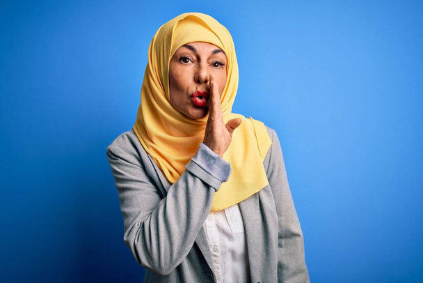 Moyen âge brunette femme d'affaires portant hijab traditionnel musulman sur fond bleu main sur la bouche racontant rumeur secrète, chuchotant conversation malicieuse - Photo, image