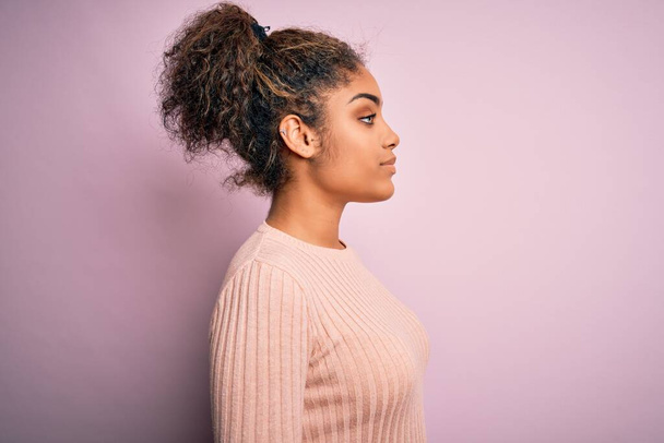 若いです美しいですアフリカ系アメリカ人の女の子身に着けていますカジュアルなセーター上にピンクの背景を見て,リラックスプロファイルポーズとともに自然な顔と自信笑顔. - 写真・画像