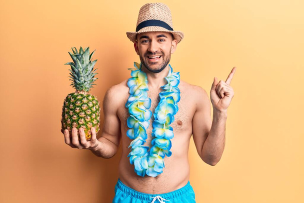 Mladý hezký plešatý muž v plavkách a hawaiian lei drží ananas s úsměvem šťastný ukazuje rukou a prstem na stranu  - Fotografie, Obrázek