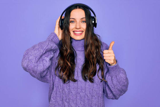 Young beautiful woman with blue eyes listening to music and dancing using headphones happy with big smile doing ok sign, thumb up with fingers, excellent sign - Valokuva, kuva