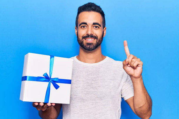 Young hispanic man holding gift smiling with an idea or question pointing finger with happy face, number one  - Photo, Image