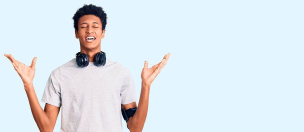 Jeune homme afro-américain portant des vêtements de gymnastique et utilisant des écouteurs célébrant fou et fou pour le succès avec les bras levés et les yeux fermés hurlant excité. concept gagnant  - Photo, image