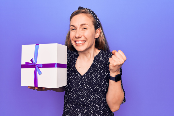 Young beautiful blonde woman holding birthday present box screaming proud, celebrating victory and success very excited with raised arm  - Foto, imagen
