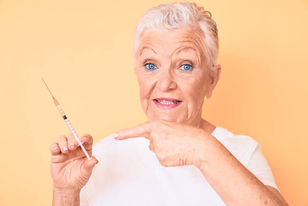 Senior όμορφη γυναίκα με μπλε μάτια και γκρι μαλλιά κρατώντας σύριγγα χαμογελώντας χαρούμενος δείχνοντας με το χέρι και το δάχτυλο  - Φωτογραφία, εικόνα