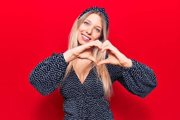 Jonge blonde vrouw draagt casual kleding glimlachend in liefde tonen hart symbool en vorm met de handen. romantisch concept.  - Foto, afbeelding