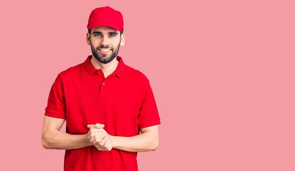 Young handsome man with beard wearing delivery uniform with hands together and crossed fingers smiling relaxed and cheerful. success and optimistic  - Photo, Image