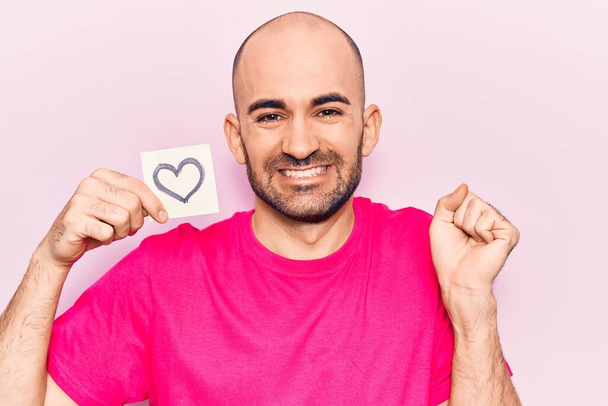 Young handsome bald man holding reminder with heart shape screaming proud, celebrating victory and success very excited with raised arm  - Photo, Image