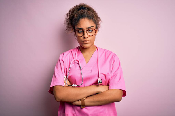 アフリカ系アメリカ人看護師の少女は、ピンクの背景に医療用の制服と聴診器を身に着けており、懐疑的で緊張しており、両腕を組んで顔の表情を否定している。負の人. - 写真・画像