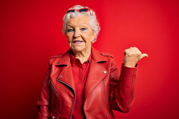 Senior kaunis harmaa tukka nainen yllään rento punainen takki ja aurinkolasit hymyillen onnellinen kasvot näköinen ja osoittaa sivuun peukalo ylöspäin. - Valokuva, kuva