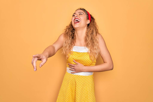 Beautiful blonde pin-up woman with blue eyes wearing diadem standing over yellow background laughing at you, pointing finger to the camera with hand over body, shame expression - Photo, Image