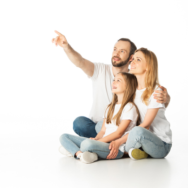 happy family sitting on floor with crossed legs while man pointing away isolated on white - Foto, immagini