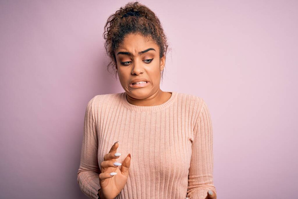 Joven chica afroamericana hermosa vistiendo suéter casual de pie sobre fondo rosa expresión disgustada, disgustada y temerosa haciendo cara de asco porque la reacción de aversión
. - Foto, imagen