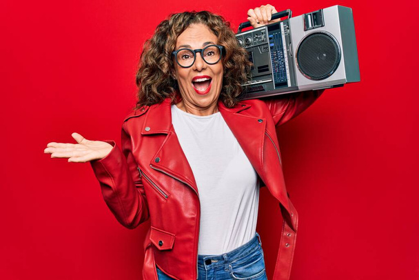 Middle age brunette hipster woman holding retro music boombox over red isolated background celebrating achievement with happy smile and winner expression with raised hand - Photo, Image