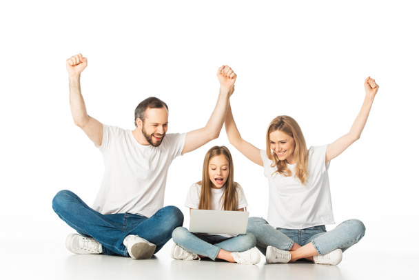 excited parents sitting on floor near happy daughter with laptop isolated on white - Photo, image