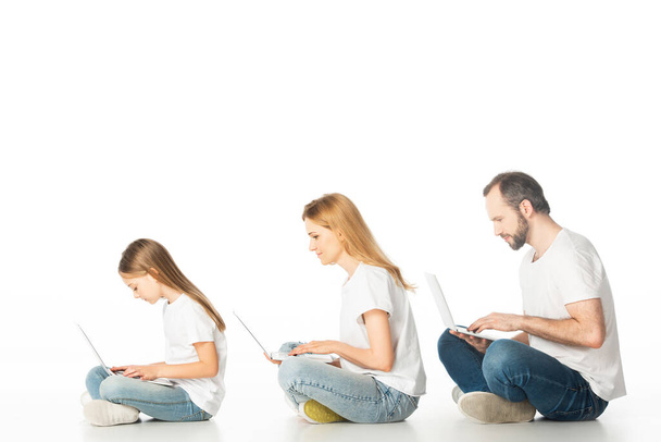 vue latérale de la famille assise sur le sol avec des ordinateurs portables sur les jambes croisées isolées sur blanc - Photo, image