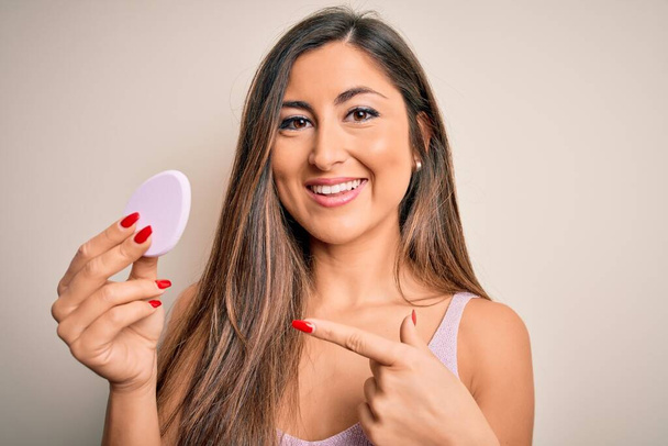 Jovem mulher bonita usando esponja de beleza com maquiagem facial sobre fundo rosa muito feliz apontando com mão e dedo
 - Foto, Imagem