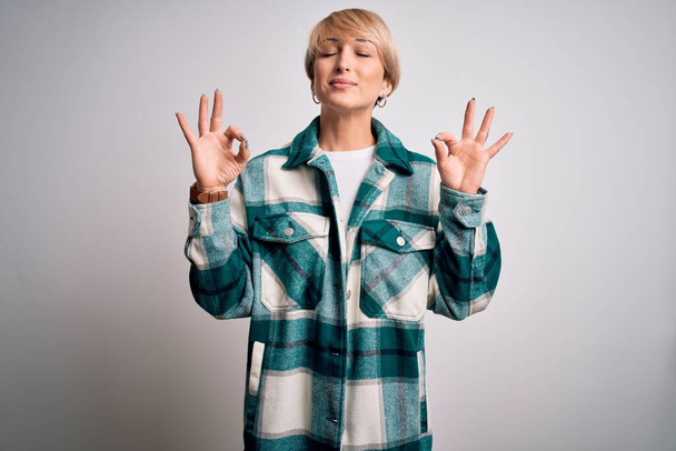 Young blonde woman with short hair wearing casual retro green shirt over isolated background relax and smiling with eyes closed doing meditation gesture with fingers. Yoga concept. - Фото, изображение