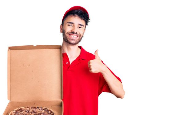 İspanyol asıllı genç adam elinde pizza kutusuyla mutlu ve pozitif gülümsüyor, başparmağıyla mükemmel ve onay işareti yapıyor.  - Fotoğraf, Görsel
