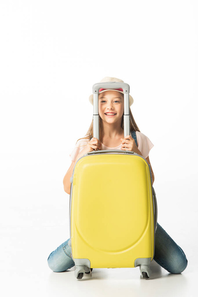 χαρούμενο κορίτσι με καπέλο κοντά σε κίτρινη ταξιδιωτική τσάντα απομονωμένη σε λευκό - Φωτογραφία, εικόνα