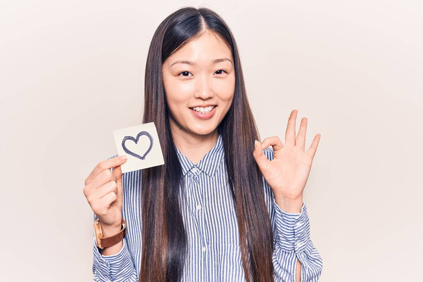 Giovane bella donna cinese che tiene il promemoria con la forma del cuore facendo segno ok con le dita, sorridente amichevole gesticolando eccellente simbolo  - Foto, immagini