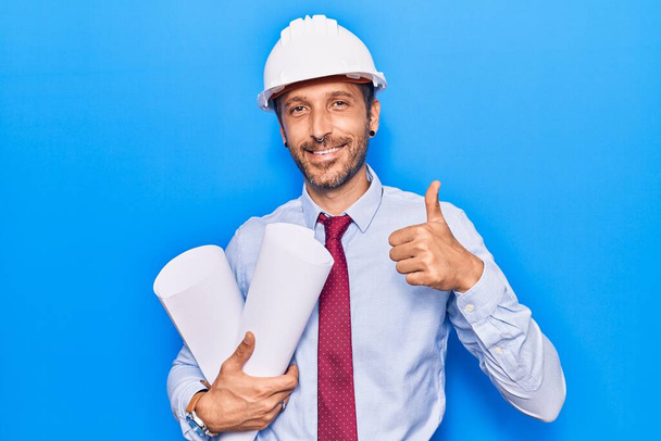 Jonge knappe man draagt architect hardhat met blauwdrukken glimlachend gelukkig en positief, duim omhoog doet uitstekend en goedkeuring teken  - Foto, afbeelding