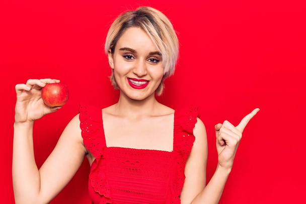 Jovem bela mulher loira segurando maçã vermelha sorrindo feliz apontando com a mão e o dedo para o lado
  - Foto, Imagem