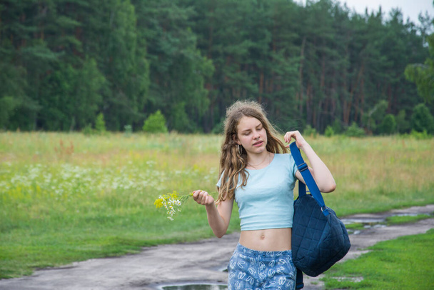 Im Sommer reist ein Teenager-Mädchen. Mit einem Rucksack auf den Schultern und einem Blumenstrauß geht sie die Straße entlang.. - Foto, Bild