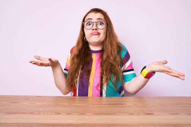 Jeune femme rousse assise sur la table portant des lunettes expression vide et confus avec les bras et les mains levées. concept de doute.  - Photo, image