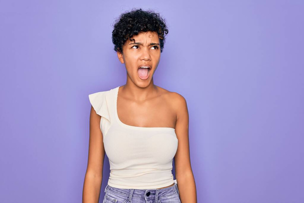 Νεαρή όμορφη Αφρικανή Αφρικάνα Αφρικάνα που φοράει casual t-shirt πάνω από μωβ φόντο θυμωμένη και θυμωμένη ουρλιάζοντας απογοητευμένη και έξαλλη, φωνάζοντας με θυμό. Οργή και επιθετική αντίληψη. - Φωτογραφία, εικόνα
