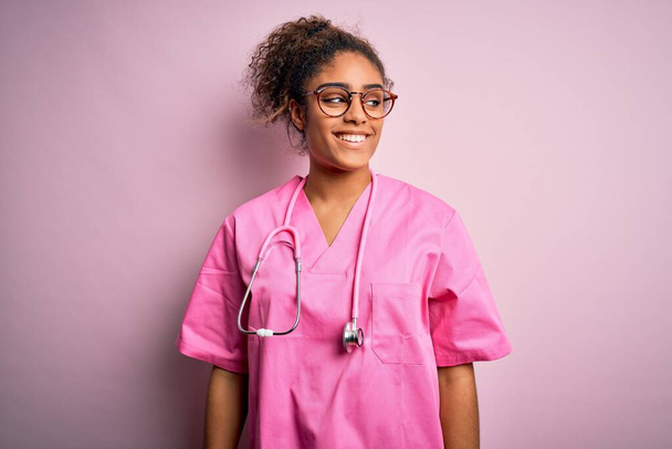 African american nurse girl wearing medical uniform and stethoscope over pink background looking away to side with smile on face, natural expression. Laughing confident. - Foto, immagini