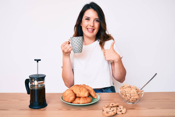 Young beautiful brunette woman sitting on the table having breakfast smiling happy and positive, thumb up doing excellent and approval sign  - Фото, изображение