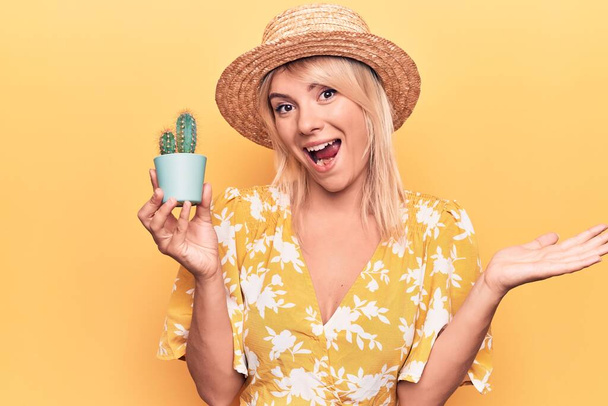 Mulher loira bonita usando chapéu de verão segurando vaso de planta de cacto sobre fundo amarelo celebrando realização com sorriso feliz e expressão vencedora com mão levantada
 - Foto, Imagem