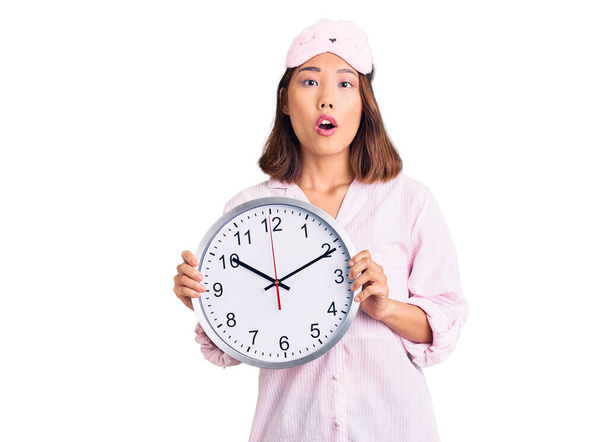 Young beautiful chinese girl wearing sleep mask and pajama holding big clock scared and amazed with open mouth for surprise, disbelief face  - Photo, Image