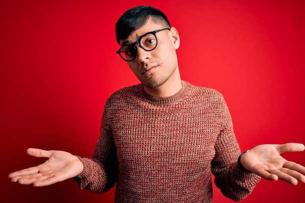 Jovem homem hispânico bonito vestindo óculos nerd sobre fundo vermelho indolente e confundido com braços abertos, nenhum conceito ideia
. - Foto, Imagem