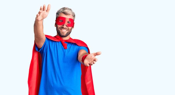 Young blond man wearing super hero custome looking at the camera smiling with open arms for hug. cheerful expression embracing happiness.  - Photo, Image