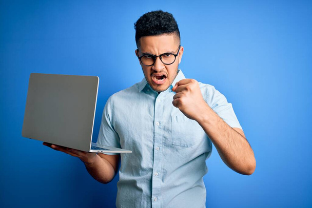 Young handsome businessman wearing glasses working using laptop over blue background annoyed and frustrated shouting with anger, crazy and yelling with raised hand, anger concept - Фото, изображение