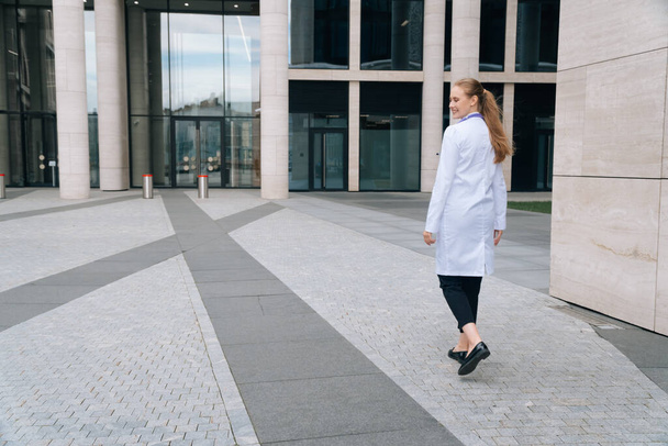 A young girl doctor in a white robe goes against the background of the medical clinic. The concept of a general plan for a doctor's work - Fotografie, Obrázek