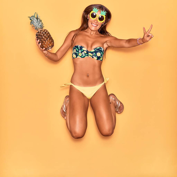 ビキニを着た若い美しいヒスパニック系の女性と面白いサングラスを笑顔幸せ。顔に笑顔でジャンプ隔離された黄色の背景にパイナップルを保持. - 写真・画像